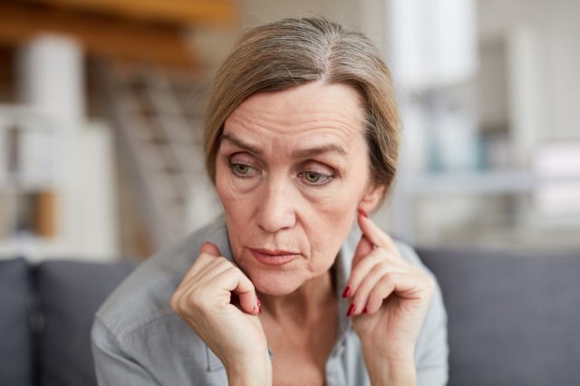 Portrait of sad mature woman sitting on sofa at home and looking away with worry and anxiety.