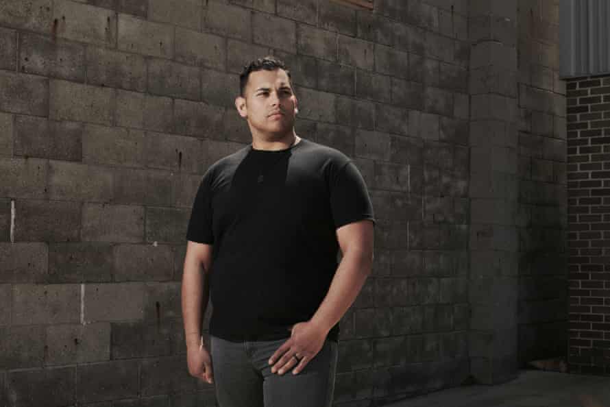 The man in a black shirt stands for a portrait in front of a concrete brick wall
